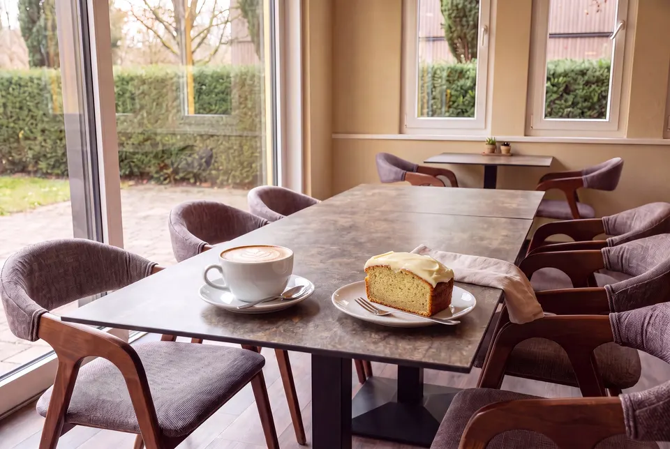 Cappuccino mit Latte Art und ein Stück Bananenkuchen mit Frischkäse-Glasur am langen Tisch vor dem Gartenfenster (Symbolbild)