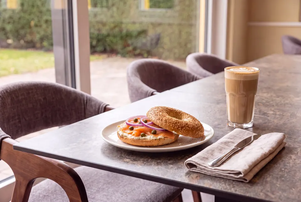 Lachsbagel mit Frischkäse, roten Zwiebeln und Kapern auf einem Teller, daneben ein Latte Macchiato, am Fenstertisch des Cafés (Symbolbild)